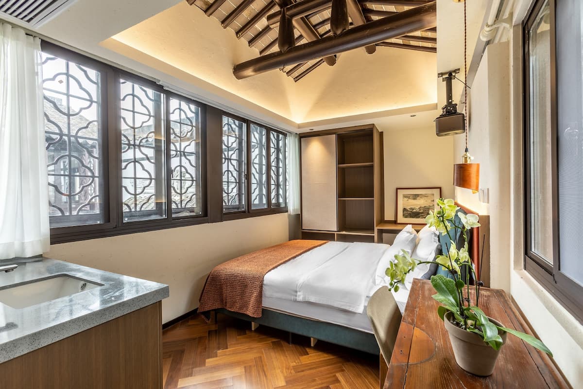 Een slaapkamer met een bed, een bureau met een wastafel, een raam met decoratieve roeders, een plafond met zichtbare balken en een potplant.