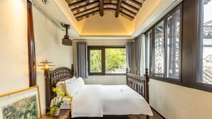 Una cama bien hecha en una habitación con suelo de madera, un ventanal con cortinas y un techo con vigas a la vista.