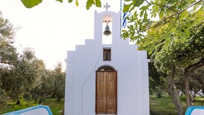 Eine kleine weiße Kirche mit einer Holztür und Stufen, die zu ihr hinaufführen, umgeben von Grünpflanzen.