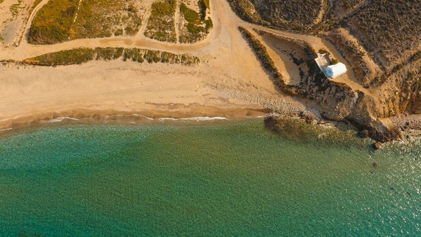 Eine Luftaufnahme einer Küstenlandschaft mit einem klaren Strand, einem kleinen Gebäude und einer felsigen Küstenlinie.