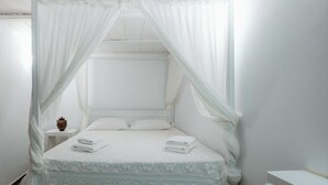 Ein weißes Himmelbett mit Spitze und Kissen, ein Nachttisch mit einer Vase sowie ein Beistelltisch mit gefalteten Handtüchern.