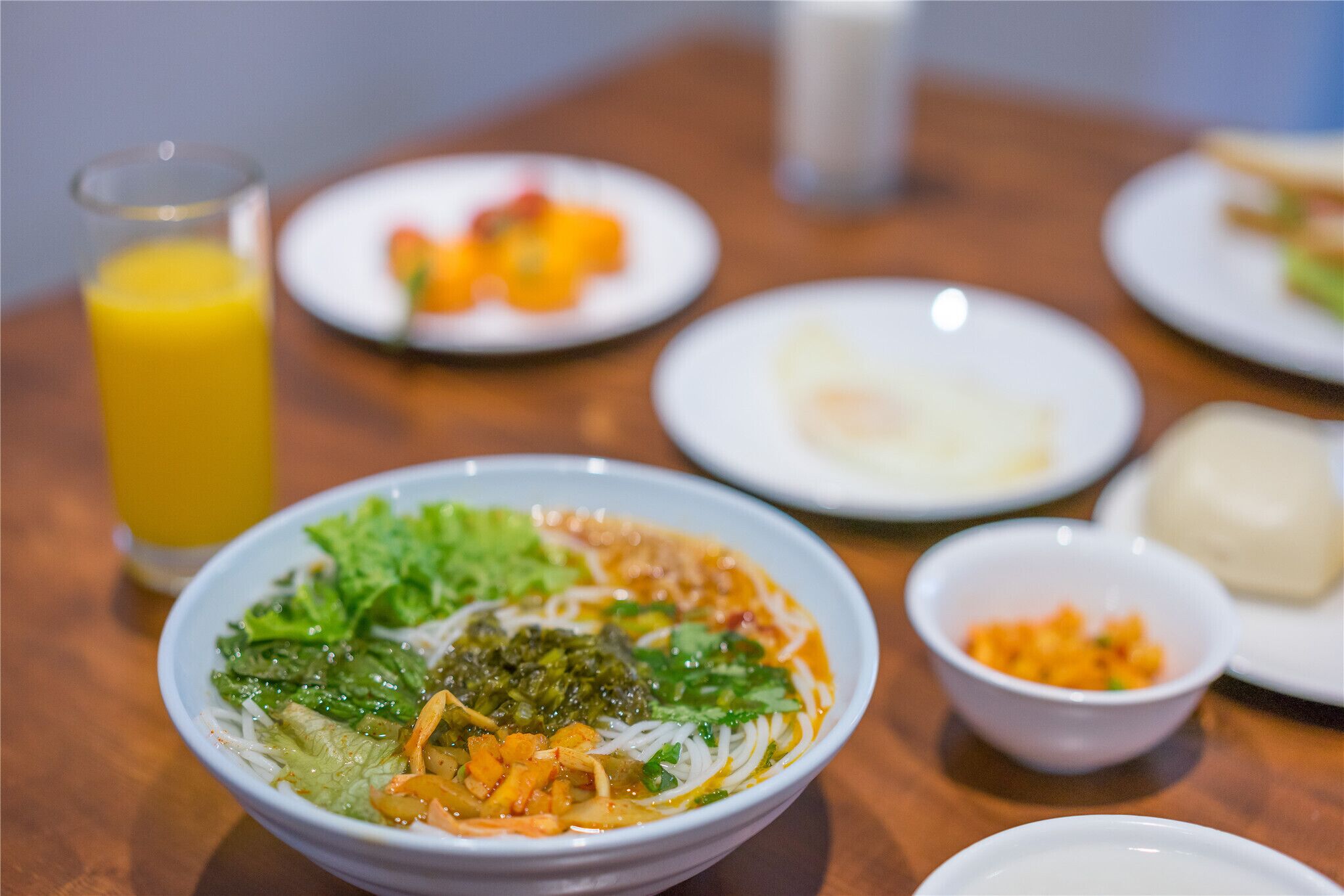 A table with a bowl of noodle soup, a glass of orange juice, and several plates of food.