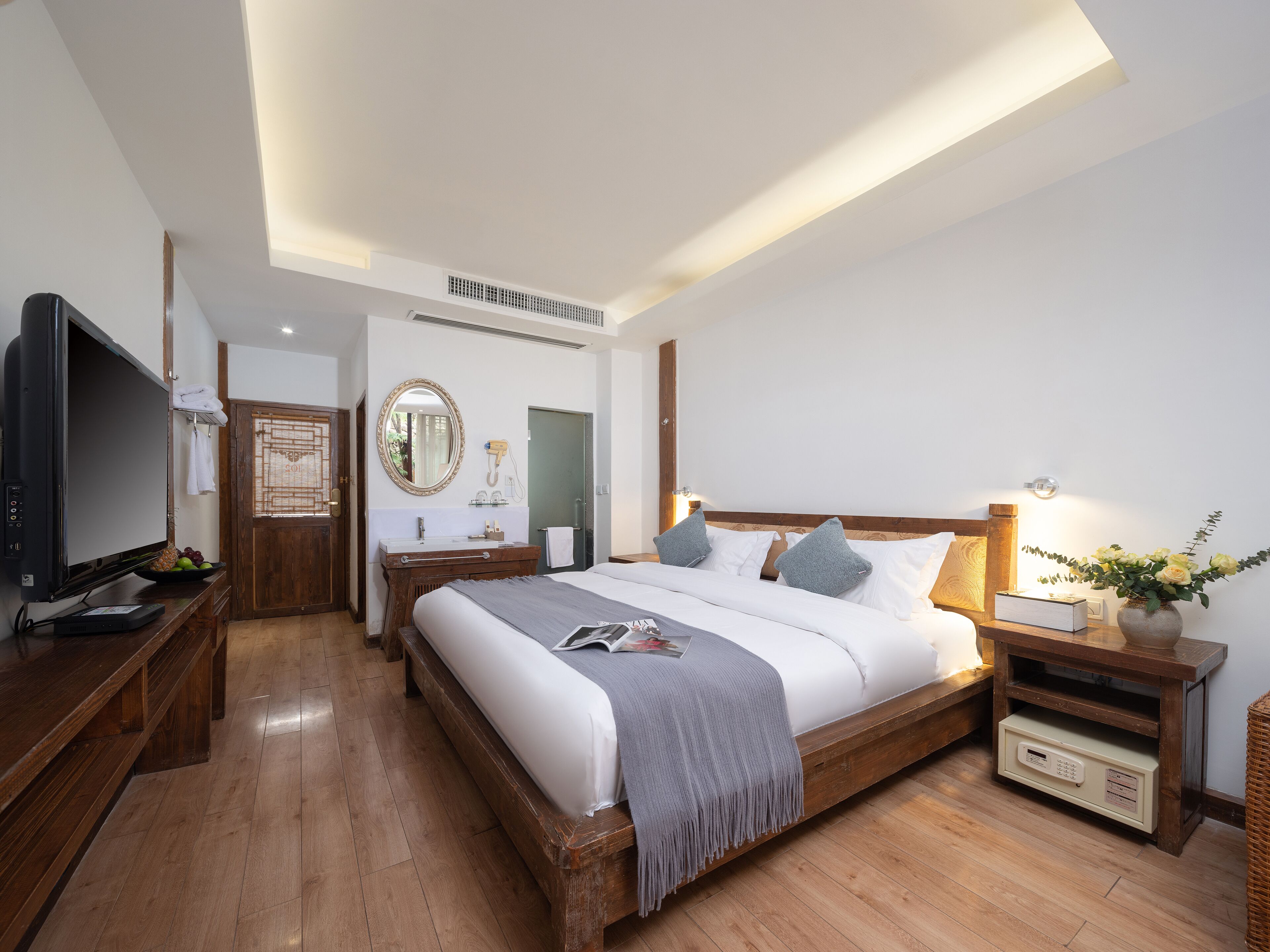 A modern hotel room with a wooden bed, a flat-screen TV, a vanity with a mirror, and a bedside table with a lamp.