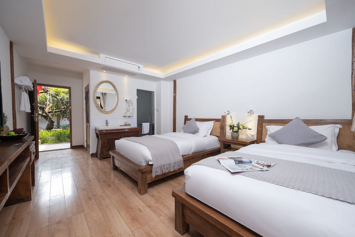 Quarto de hotel moderno com duas camas, um banco de madeira, uma penteadeira com espelho e vista para o verde lá fora.