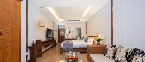 Quarto de hotel moderno com uma mesa de centro de vidro, um sofá, uma cama, uma televisão e um banheiro visível ao fundo.