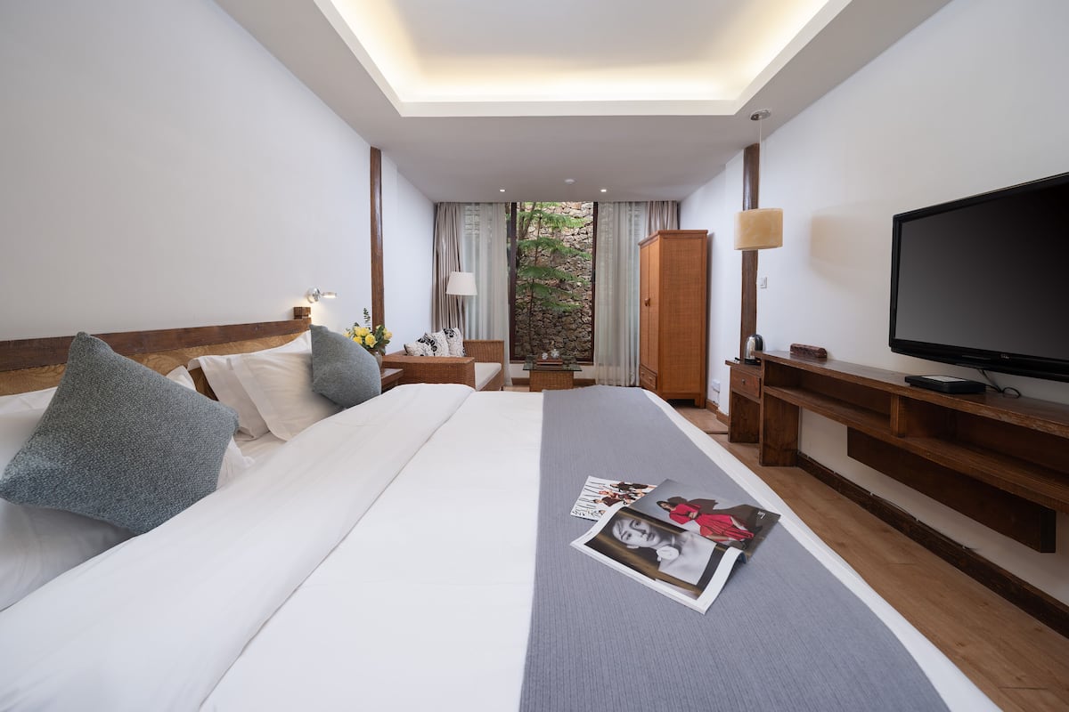 Quarto de hotel com uma cama grande, televisão, escrivaninha de madeira e vista para o verde lá fora.