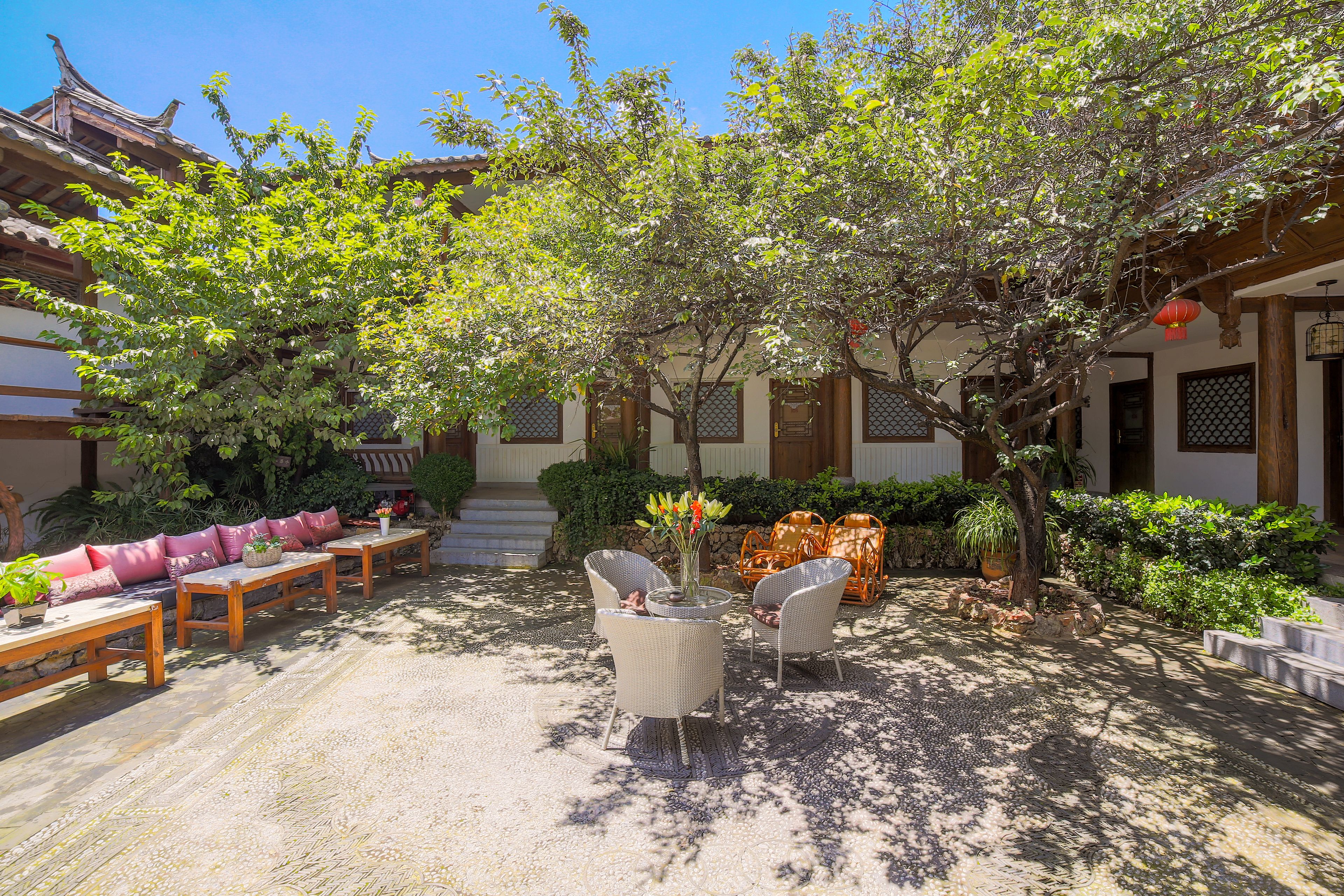 A traditional courtyard with wooden furniture, a central table with a vase, and a tree providing shade.