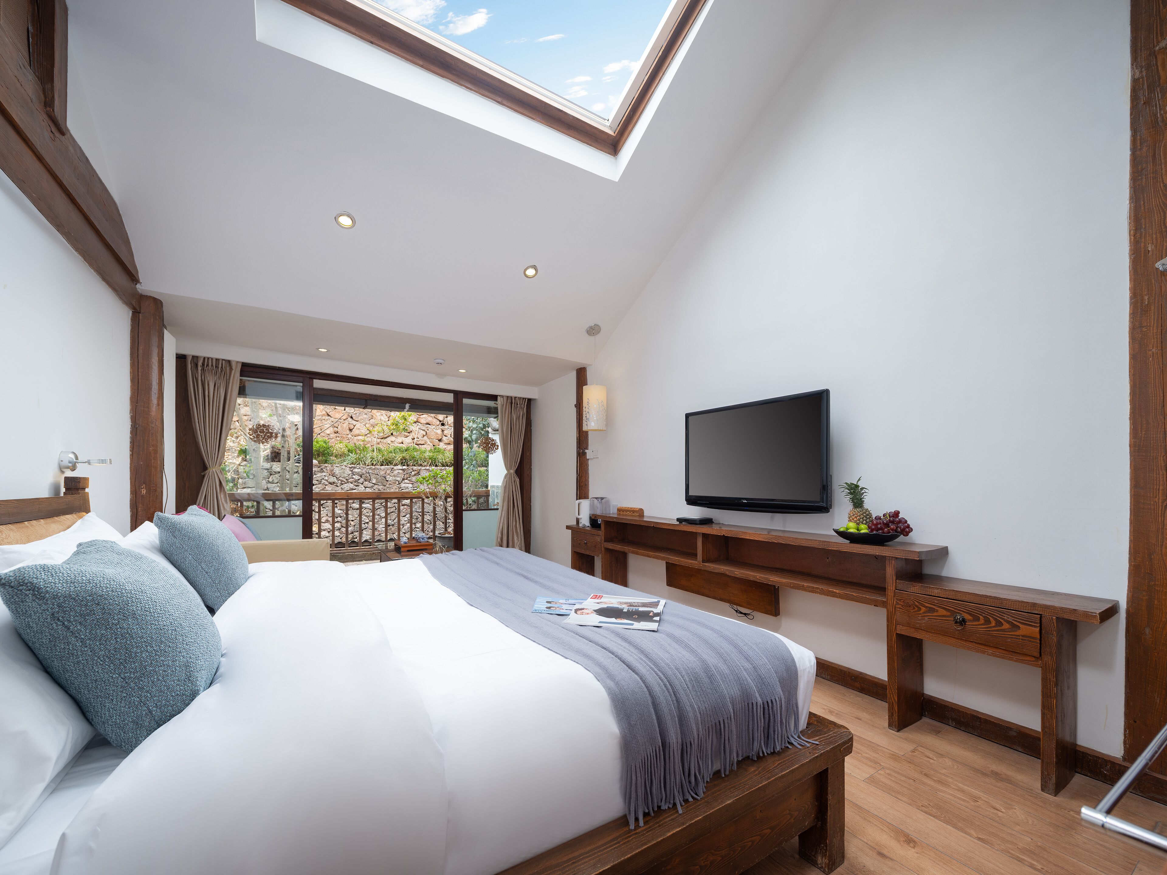 A bedroom with a large bed, a TV, a wooden desk, and a balcony with a stone wall and plants.