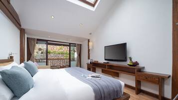 A bedroom with a large bed, a TV, a wooden desk, and a balcony with a stone wall and plants.