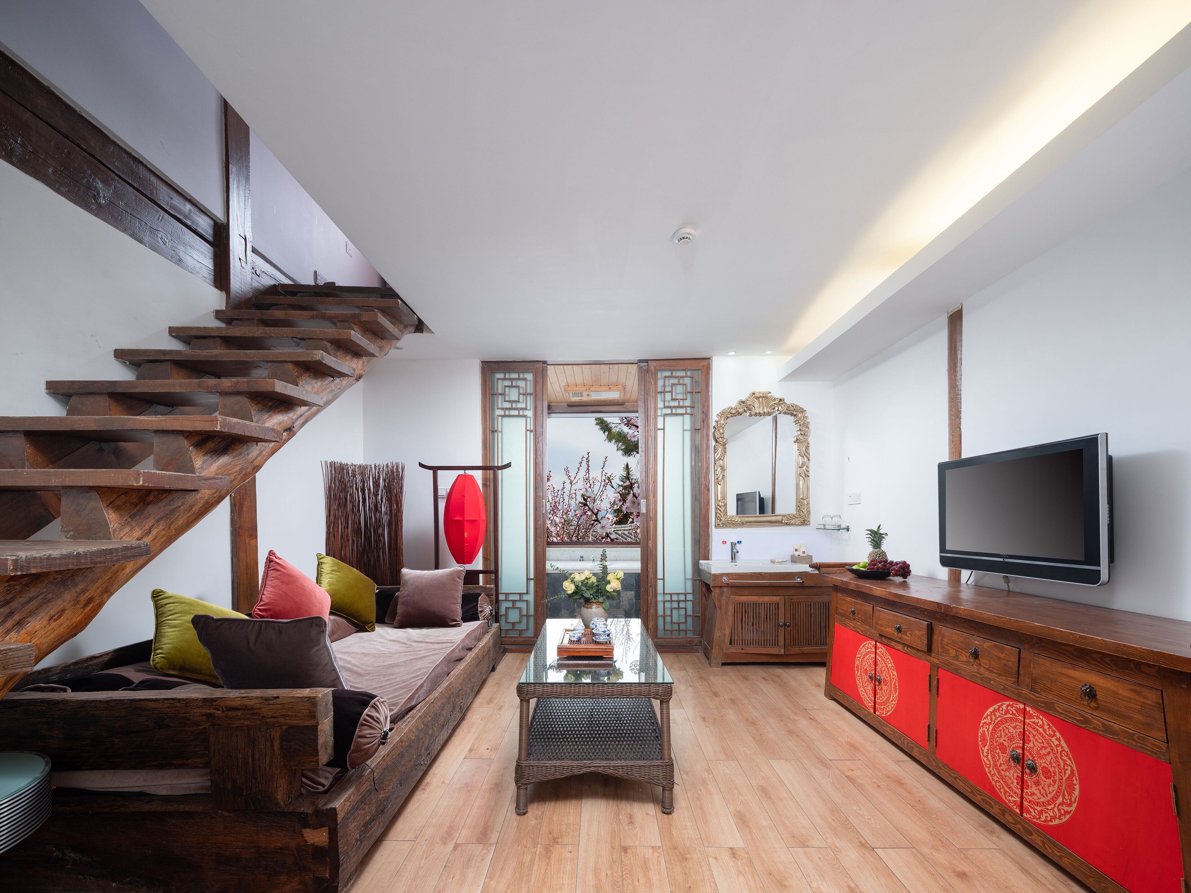 A modern living room with a wooden staircase, a sofa with cushions, a glass coffee table, a red cabinet, and a flat-screen TV.