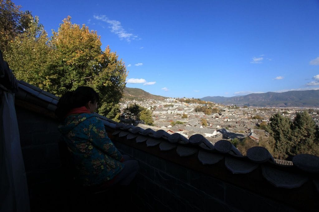 Eine Person sitzt auf einem Balkon mit Blick auf eine Stadtlandschaft, im Hintergrund sind Berge zu sehen.