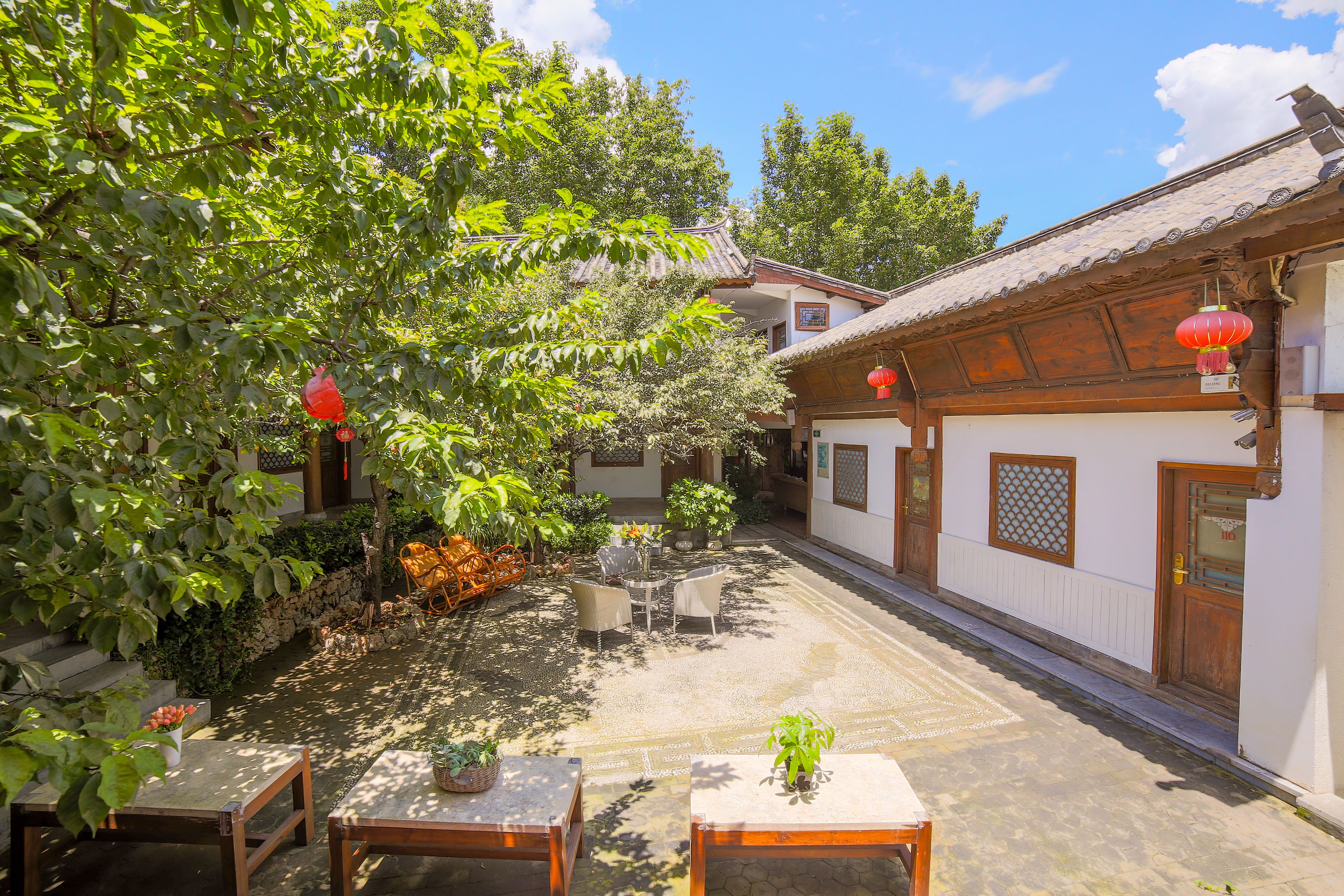 A courtyard with traditional architecture, including wooden doors and tiled roofs. There are tables and chairs arranged for outdoor dining, surrounded by lush greenery and potted plants.