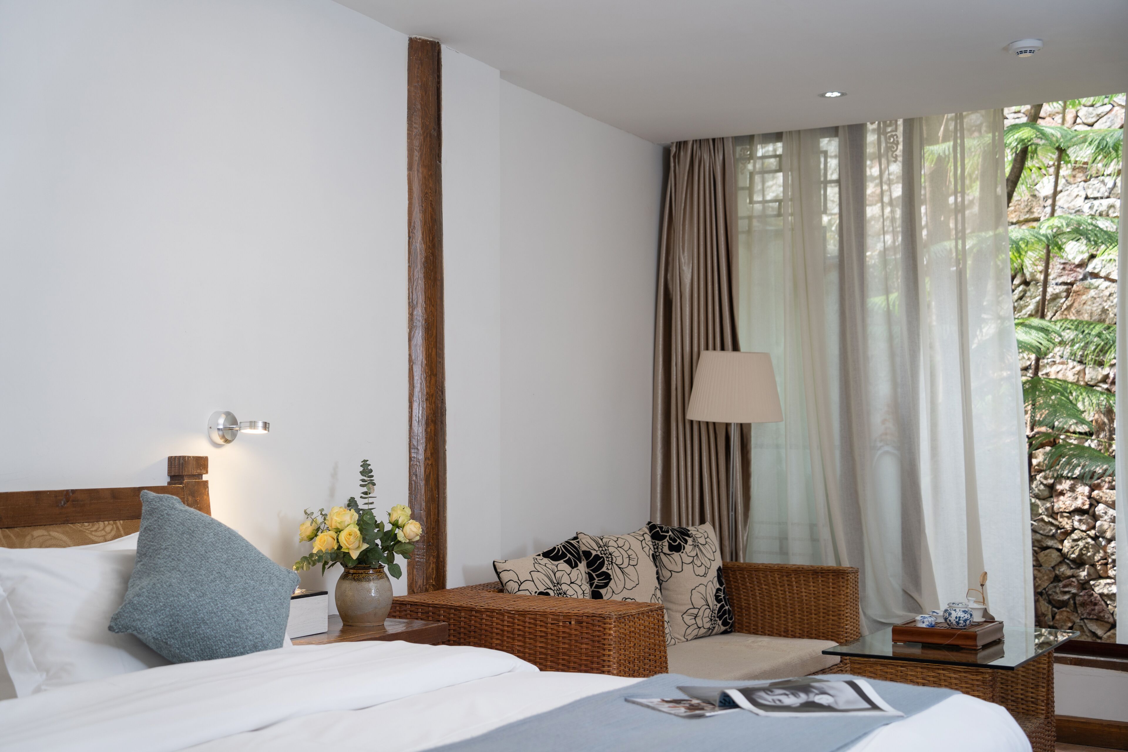 A hotel room with a bed, a wicker chair, a nightstand with a lamp, and a view of a rocky garden through sheer curtains.
