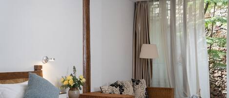 A hotel room with a bed, a wicker chair, a nightstand with a lamp, and a view of a rocky garden through sheer curtains.