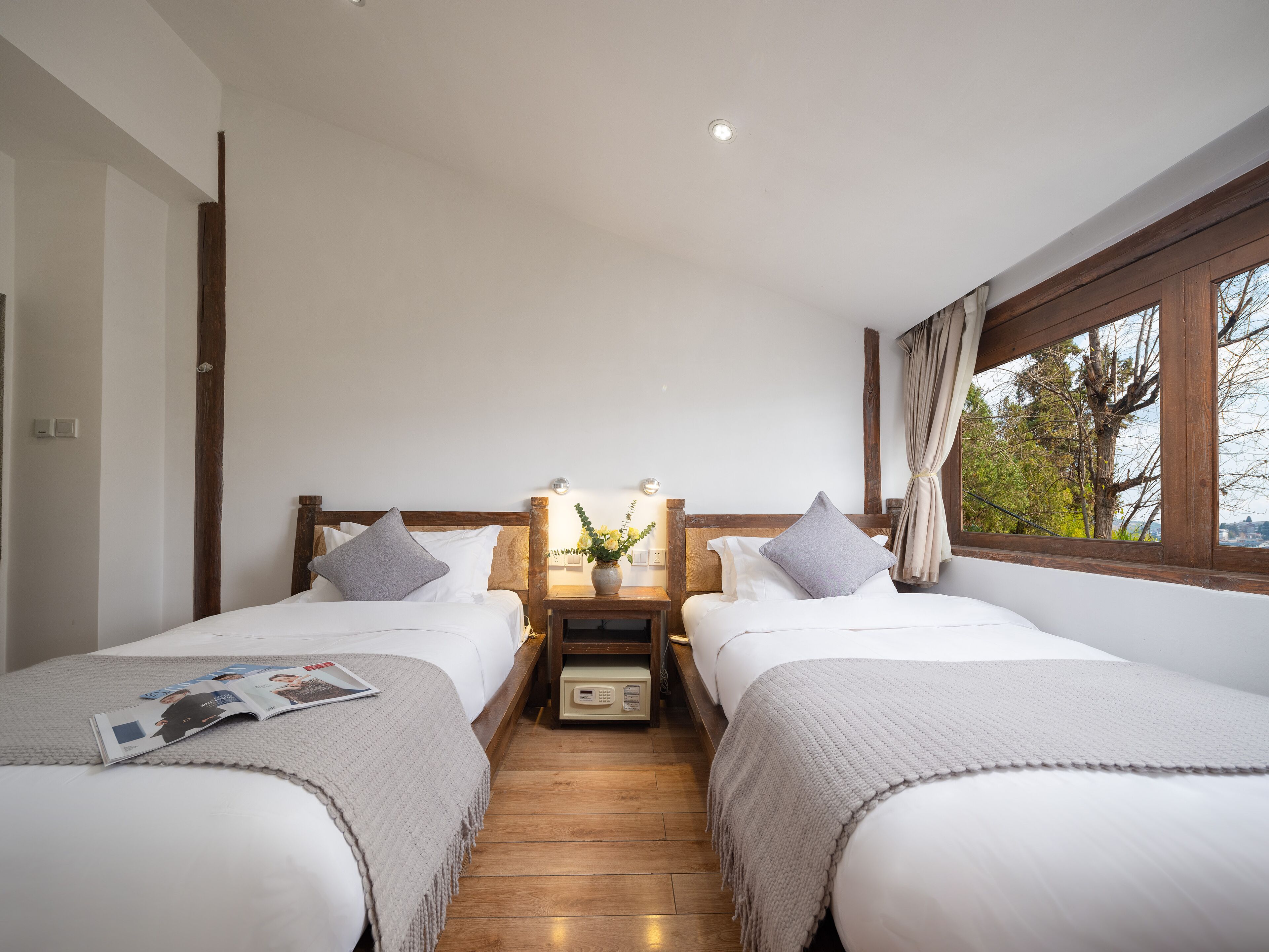 A double room with two beds, wooden headboards, a nightstand with a lamp, a small table with a vase, and a window with a view of trees.