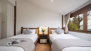 A double room with two beds, wooden headboards, a nightstand with a lamp, a small table with a vase, and a window with a view of trees.