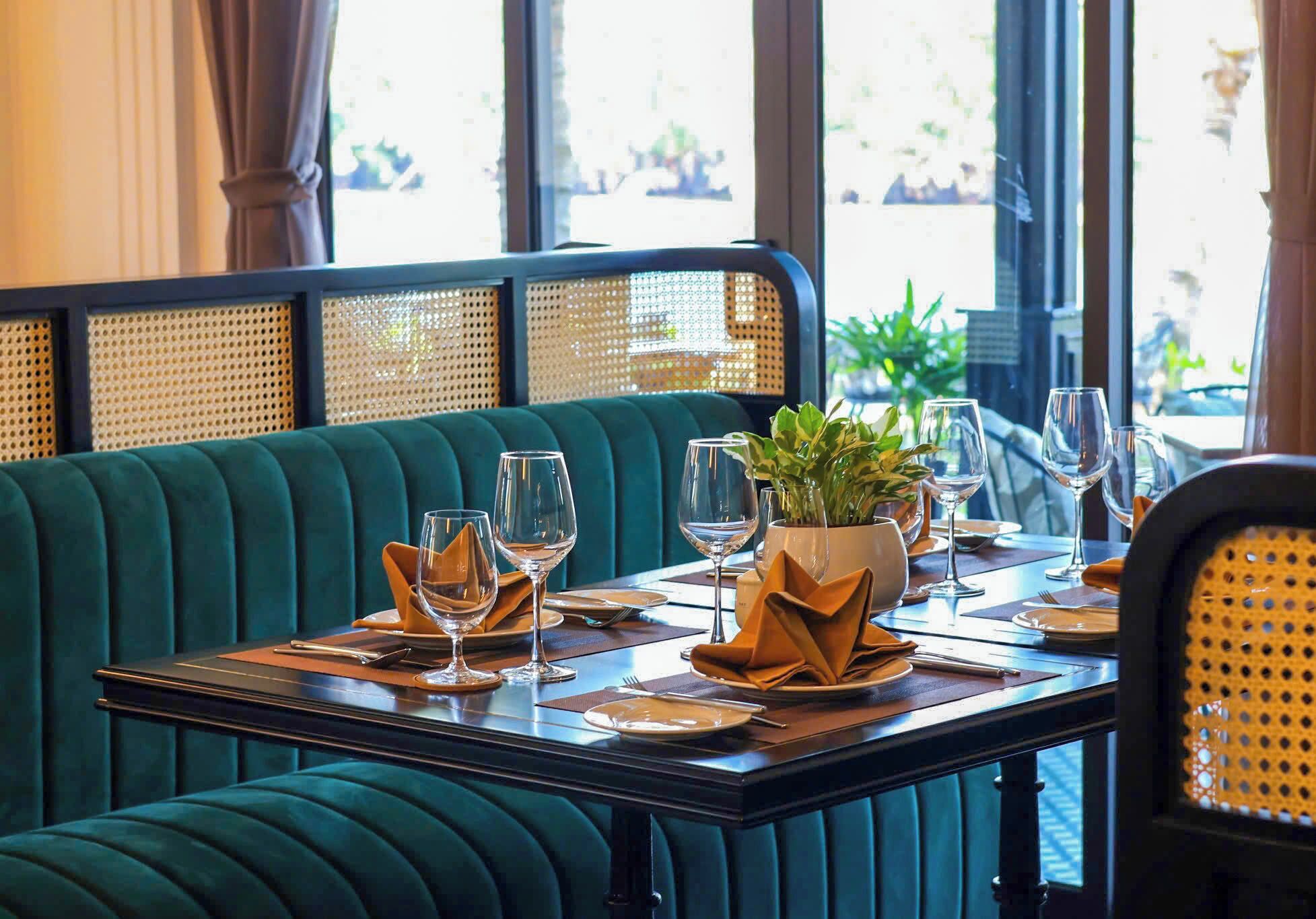 Un restaurante con un asiento tapizado en verde, una mesa dispuesta con copas de vino, platos y una planta en maceta.
