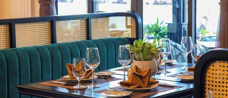 Un restaurante con un asiento tapizado en verde, una mesa dispuesta con copas de vino, platos y una planta en maceta.