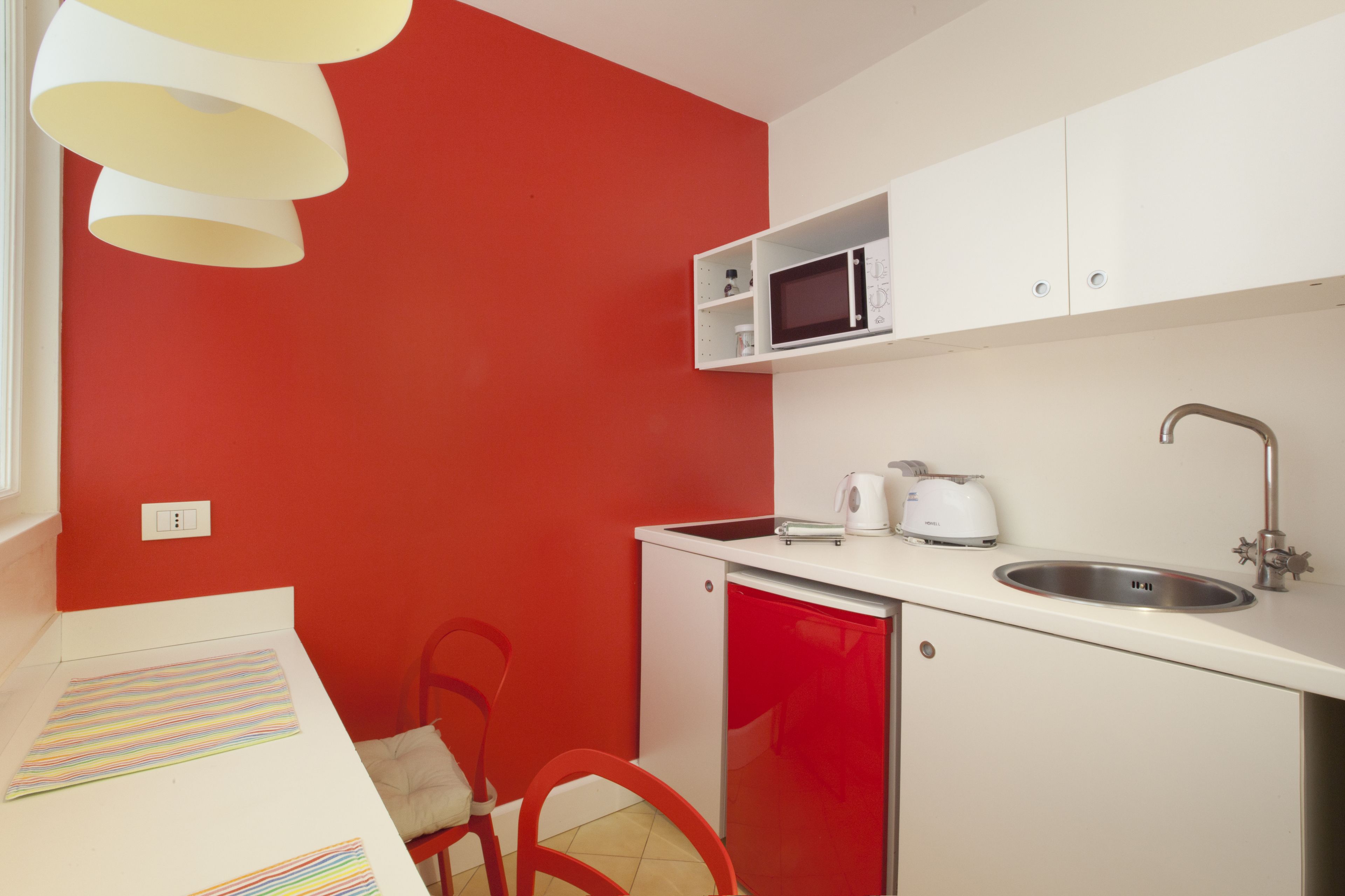 Una cocina compacta con una pared roja, armarios blancos, microondas, pileta y un refrigerador pequeño.