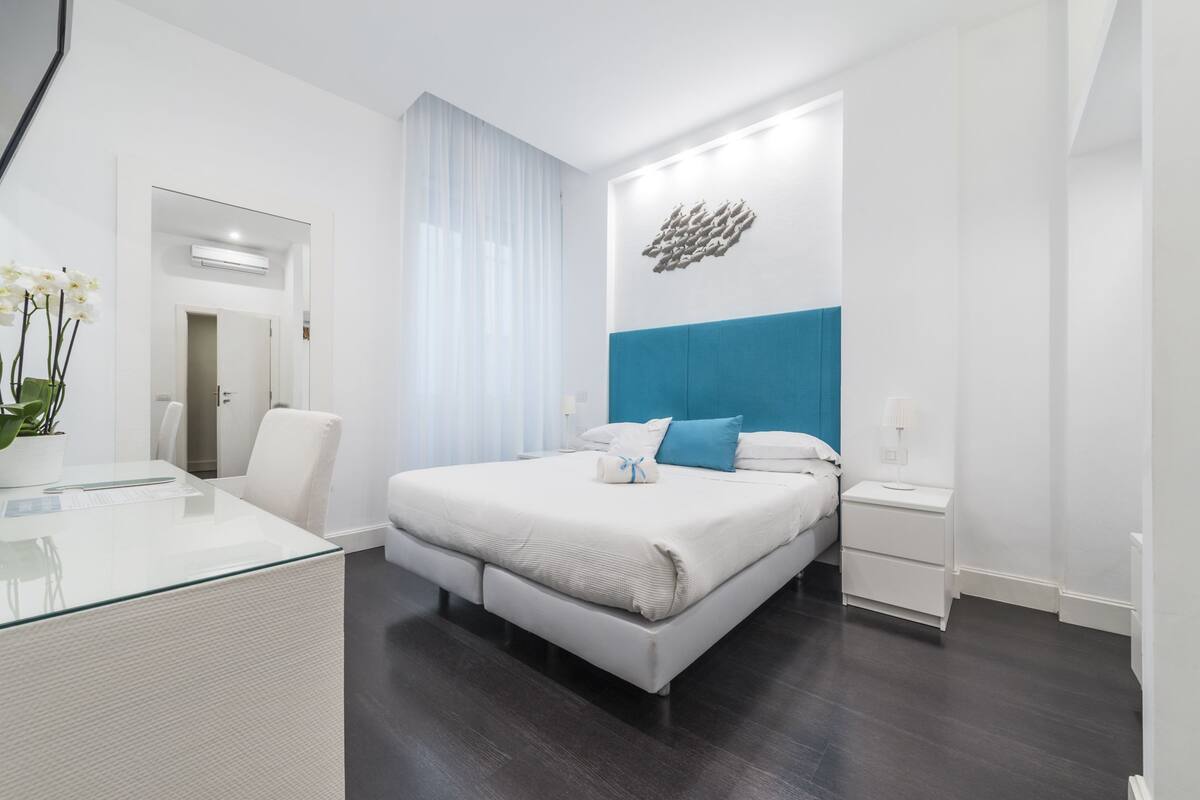 A modern hotel room with a large bed, a glass desk, a nightstand, and a wall-mounted light fixture.