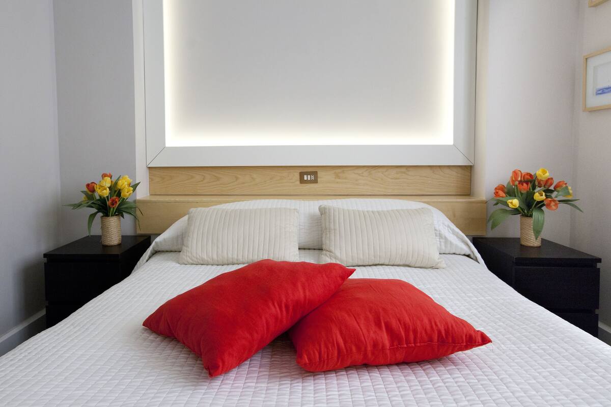A neatly made bed with white bedding and two red pillows, flanked by black nightstands with vases of flowers.