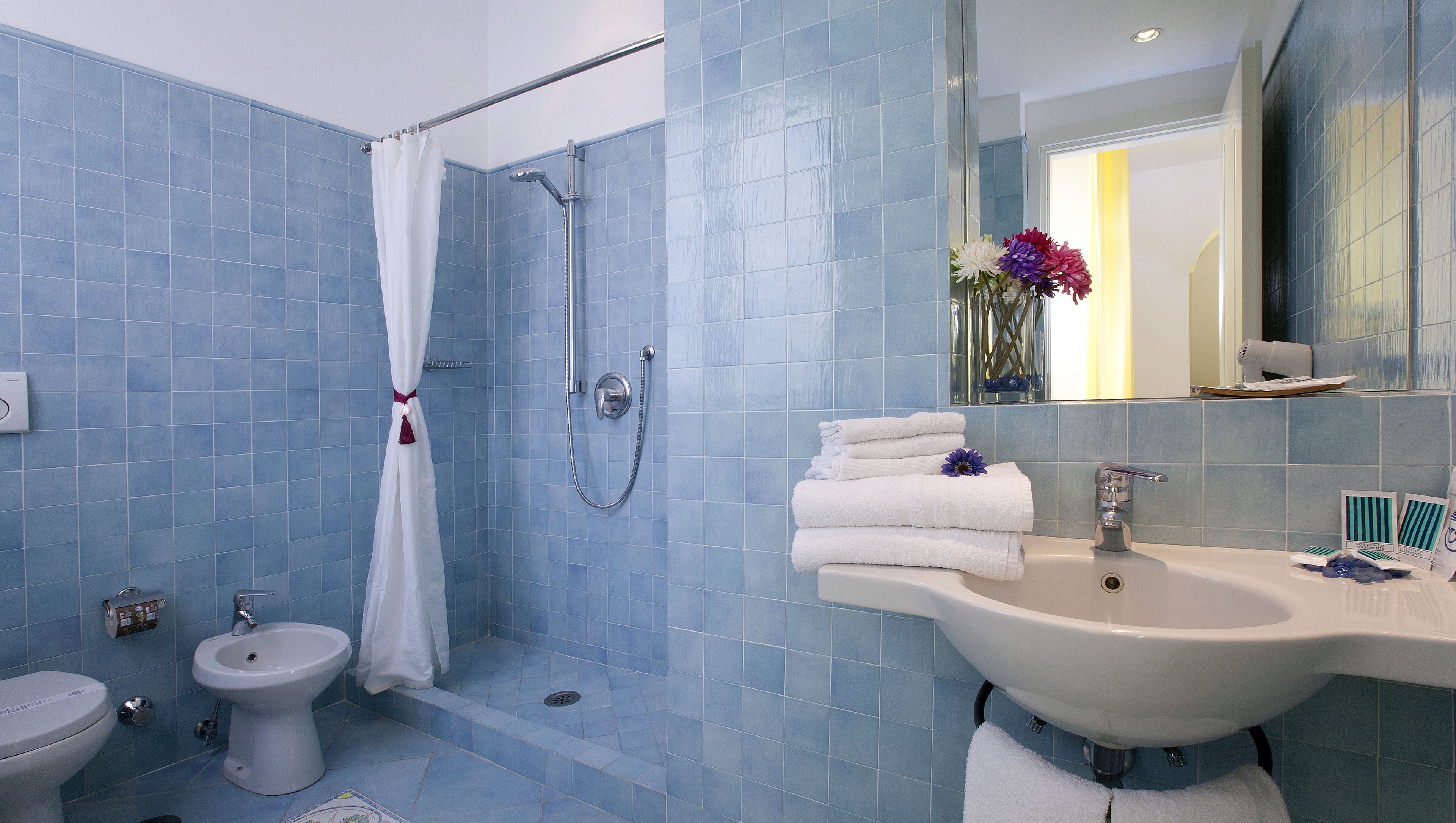 A bathroom with blue tiles, a shower, a bidet, and a sink with stacked towels.