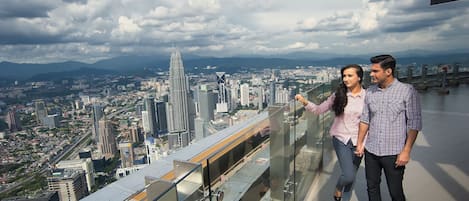 Un couple se tient sur le balcon d’un immeuble de grande hauteur, d’où l’on peut admirer un paysage urbain avec des bâtiments modernes et un gratte-ciel imposant.