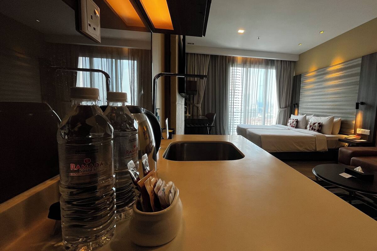 Une chambre d’hôtel avec un lit, un canapé, un bureau avec une lampe et une télévision. Il y a des bouteilles d’eau sur le bureau.