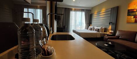 A hotel room with a bed, a sofa, a desk with a lamp, and a TV. There are water bottles on the desk.