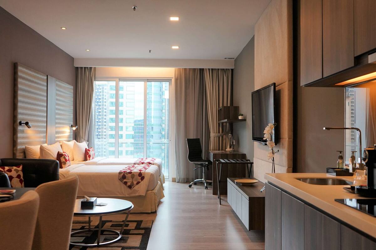 Une chambre d’hôtel comprenant un lit, un bureau, une chaise, une petite table, un téléviseur et une fenêtre avec des rideaux.