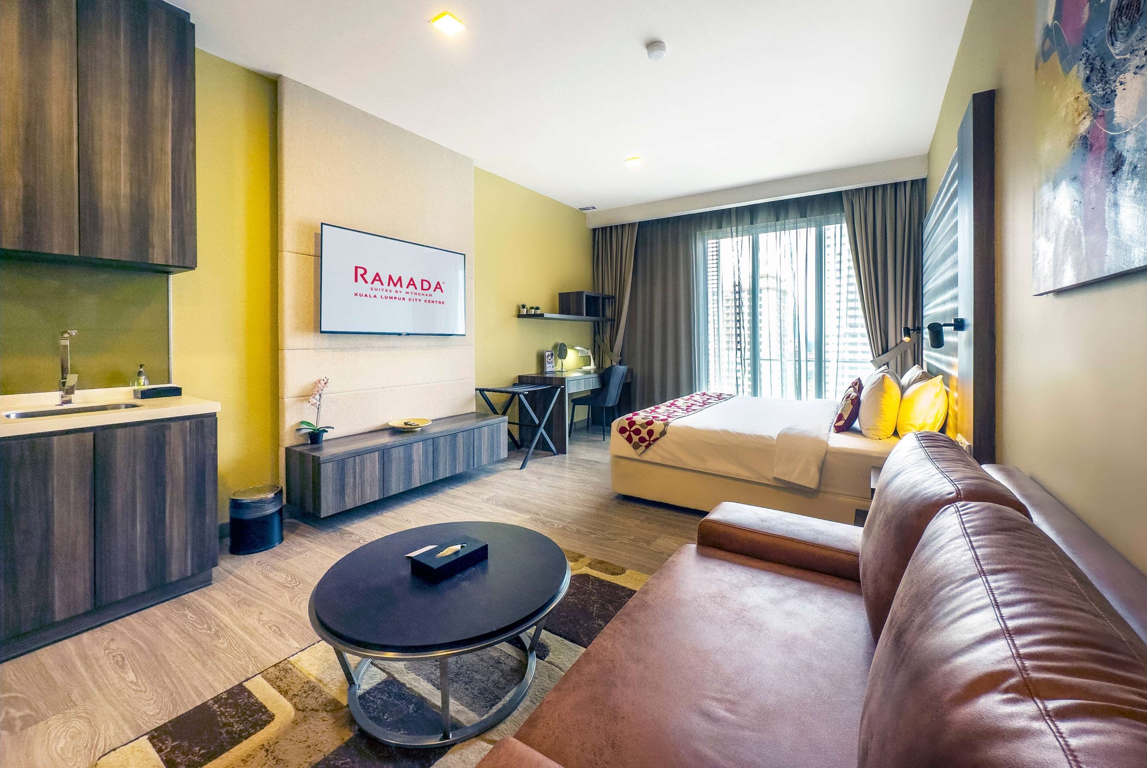A modern hotel room with a sofa, a dining table, a small kitchen area, a bed with a view, and a mounted sign.