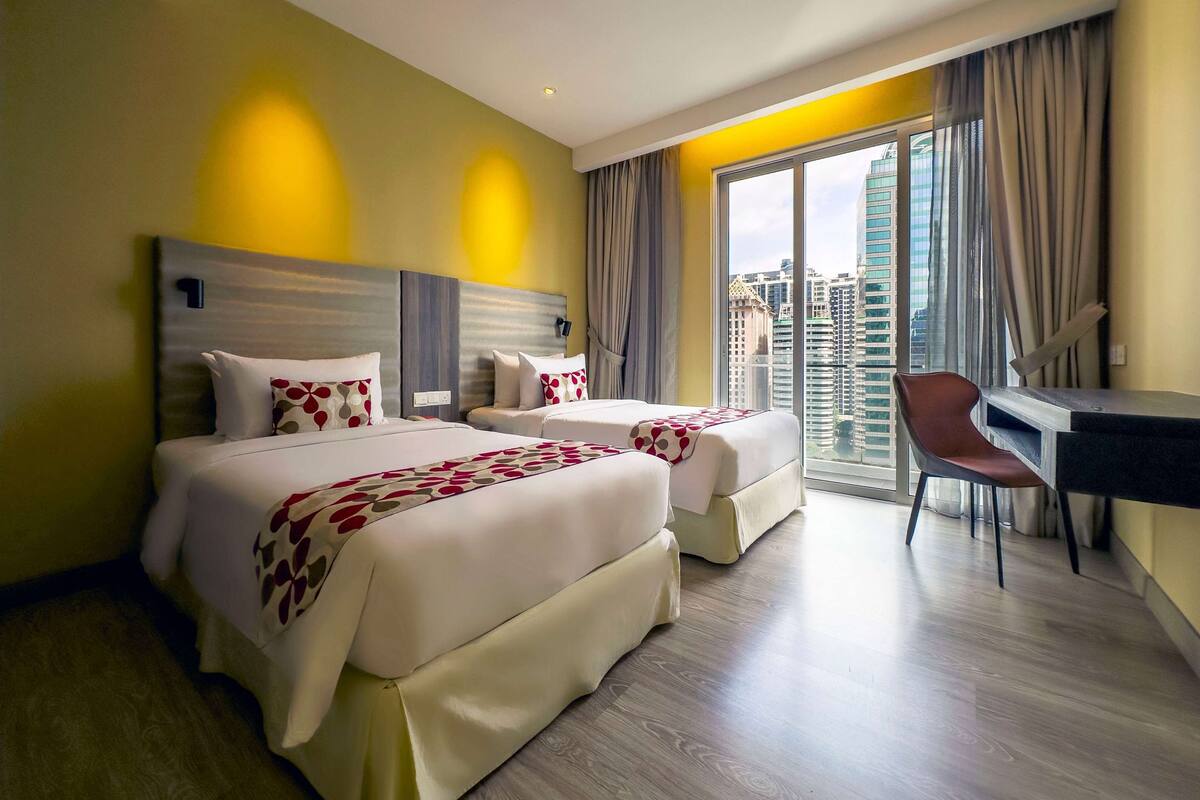 Une chambre d’hôtel avec deux lits, un bureau et une chaise. La chambre dispose d’une grande fenêtre donnant sur la ville.