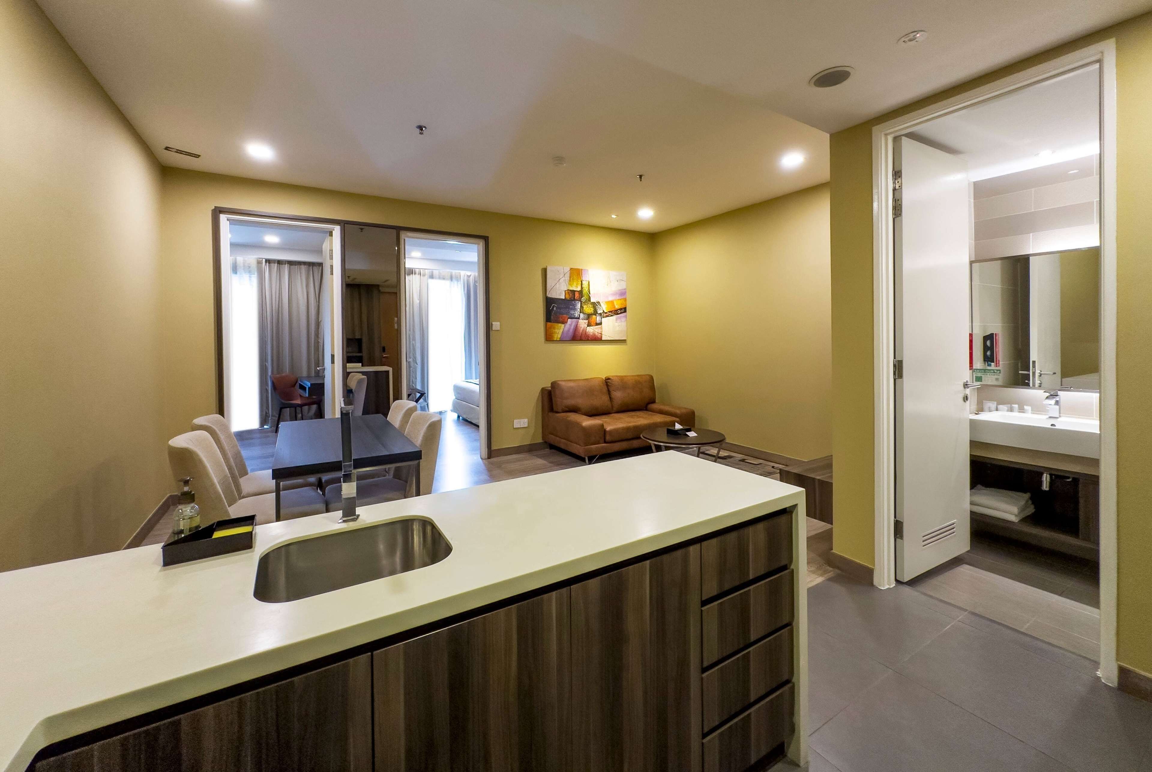 A modern kitchen with a central island, a dining area with a table and chairs, a living area with a sofa, and a bathroom with a sink and mirror.