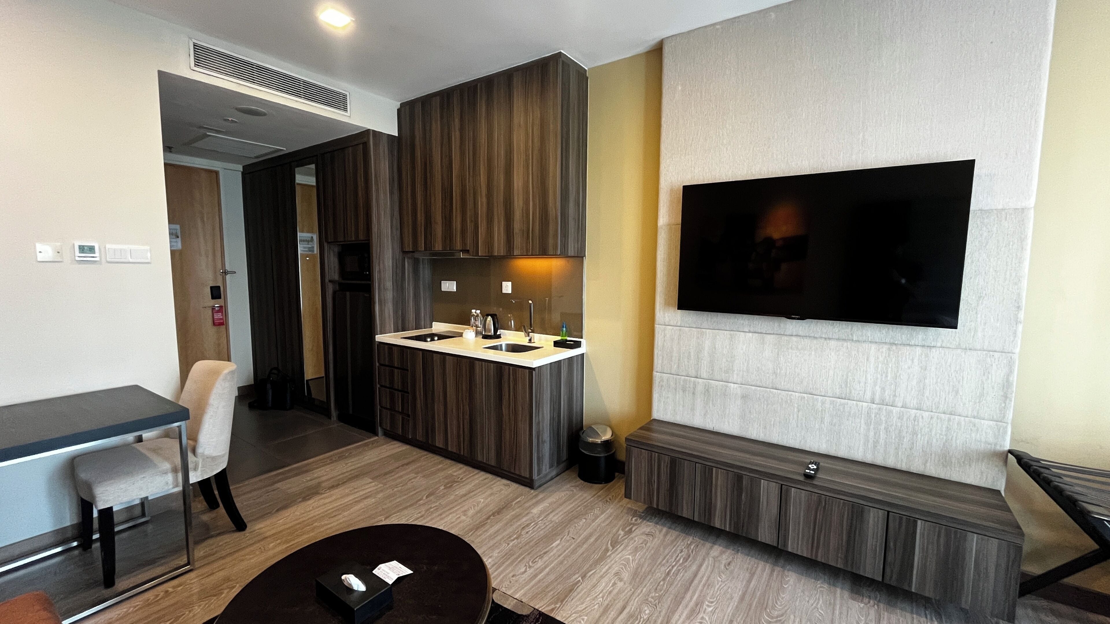 A modern hotel room with a kitchenette, a flat-screen TV mounted on the wall, a seating area with a table and chairs, and a small sofa.