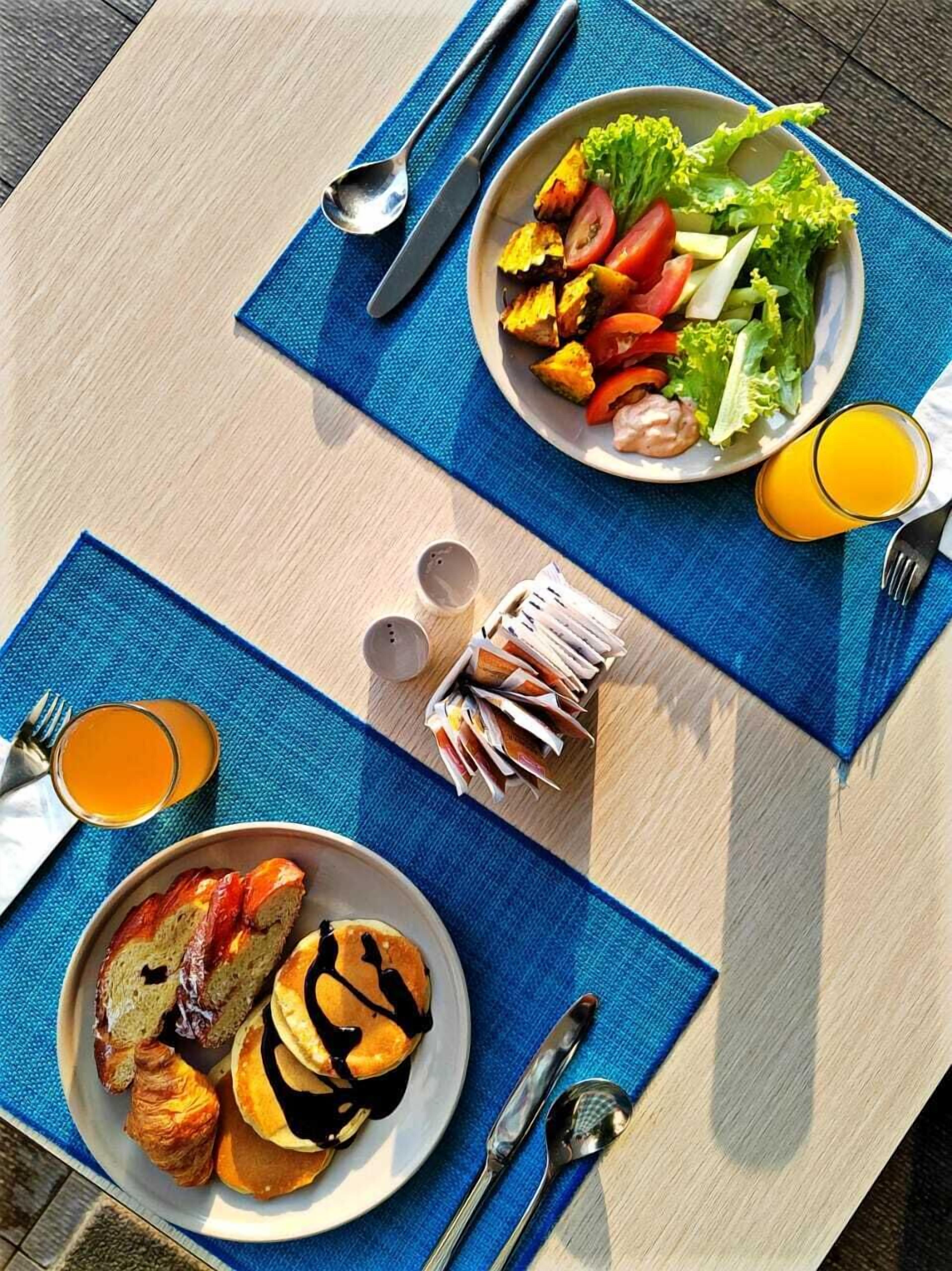 Un desayuno que incluye tostadas, cruasanes y tortitas, un bol de ensalada con vegetales y un vaso de zumo de naranja.