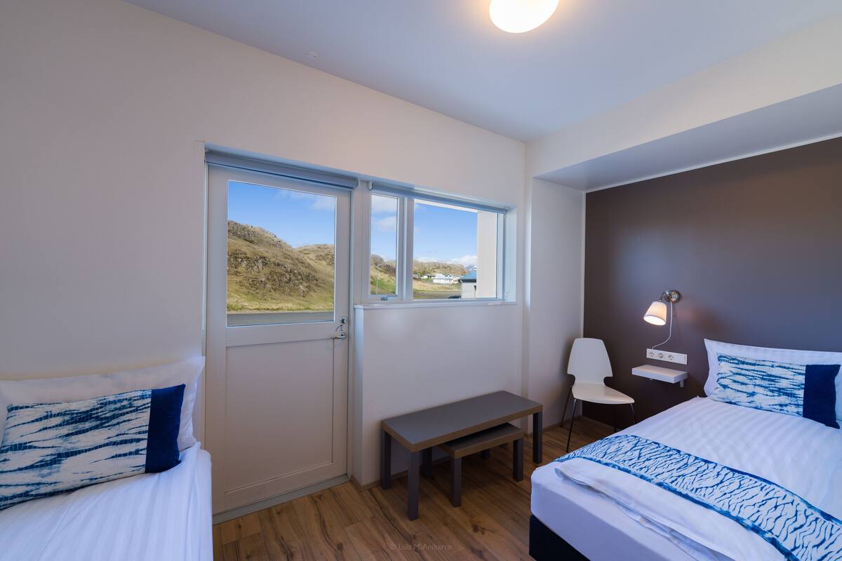 Une chambre avec deux lits, un banc, un bureau, une chaise et une fenêtre donnant sur des collines.