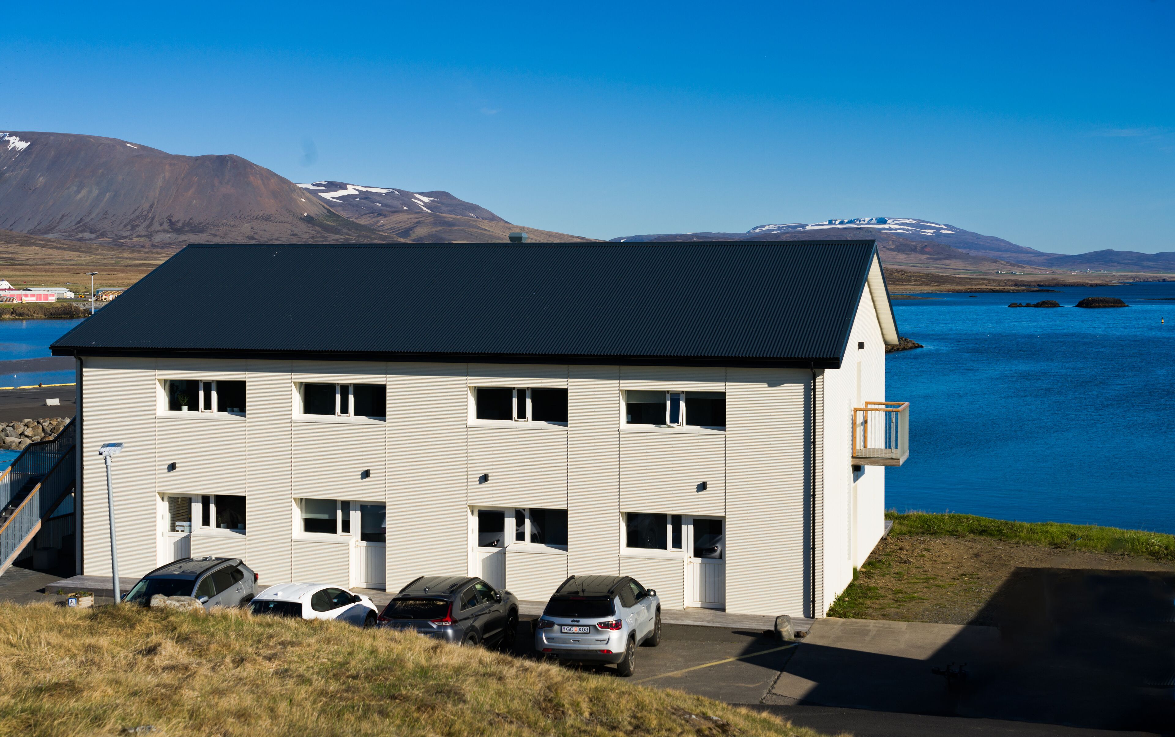 A white building with a dark roof, multiple windows, and a parking area with cars.
