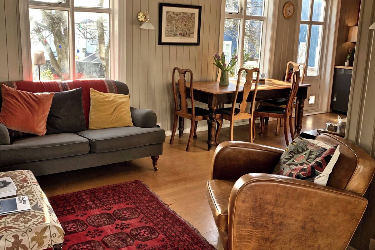 A cozy living room with a sofa, armchair, and a dining table with chairs. There are large windows allowing natural light and a view of the outside.