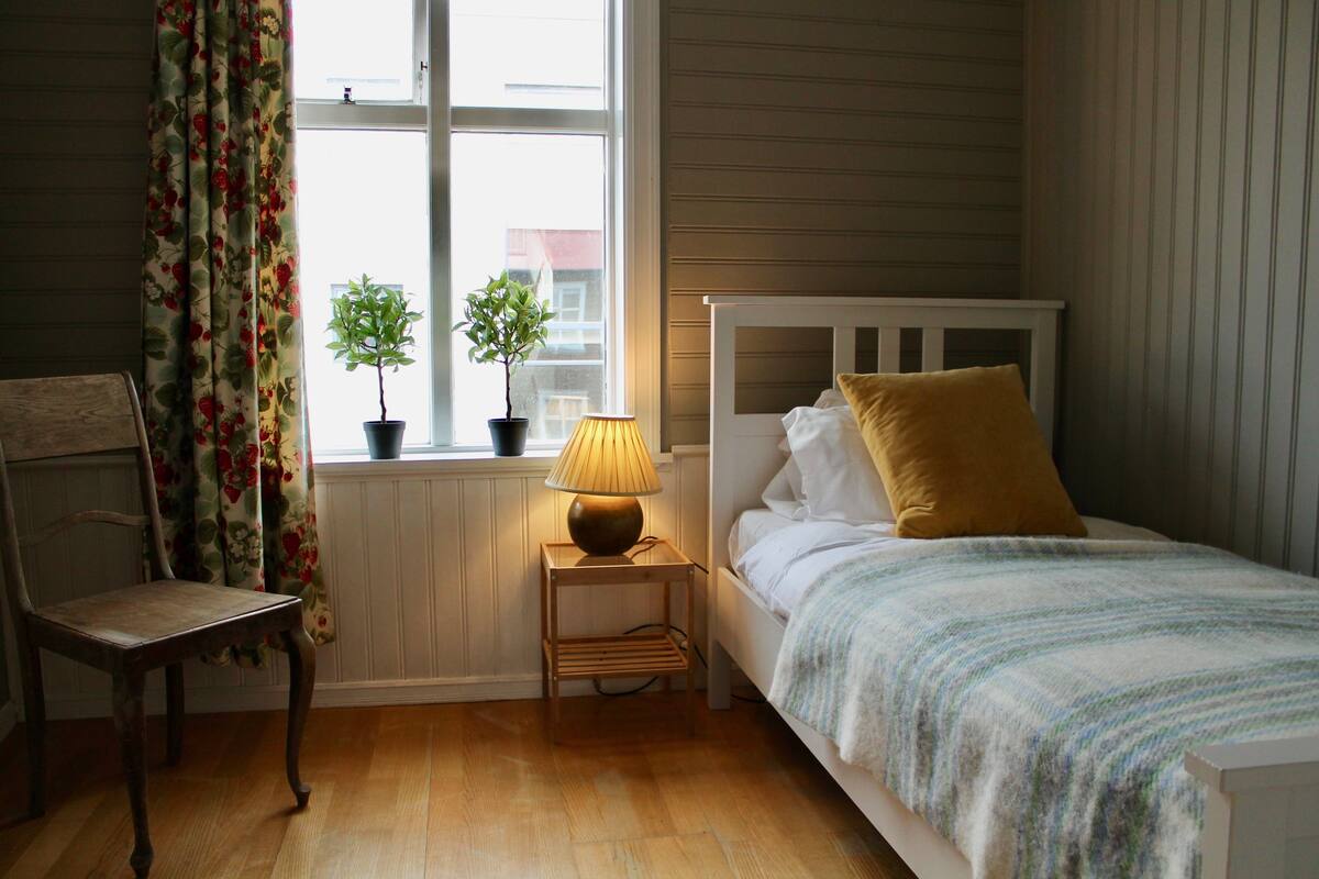 A bedroom with a wooden floor, a bed with a white headboard, a small bedside table with a lamp, a chair, and a window with curtains.
