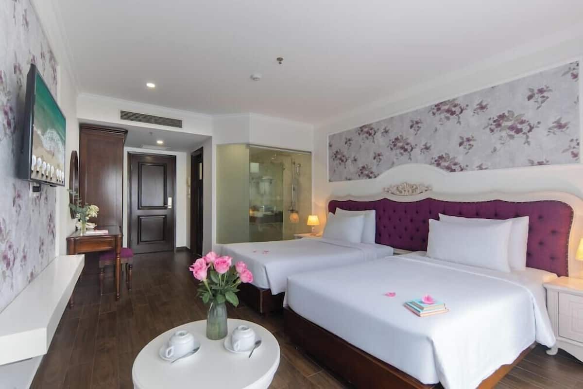 A hotel room with two beds, a sitting area with a table, and a bathroom visible through a glass door.