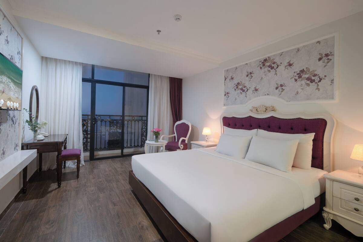 A hotel room with a large bed, a desk with a chair, a small table with a lamp, and a balcony with a railing.