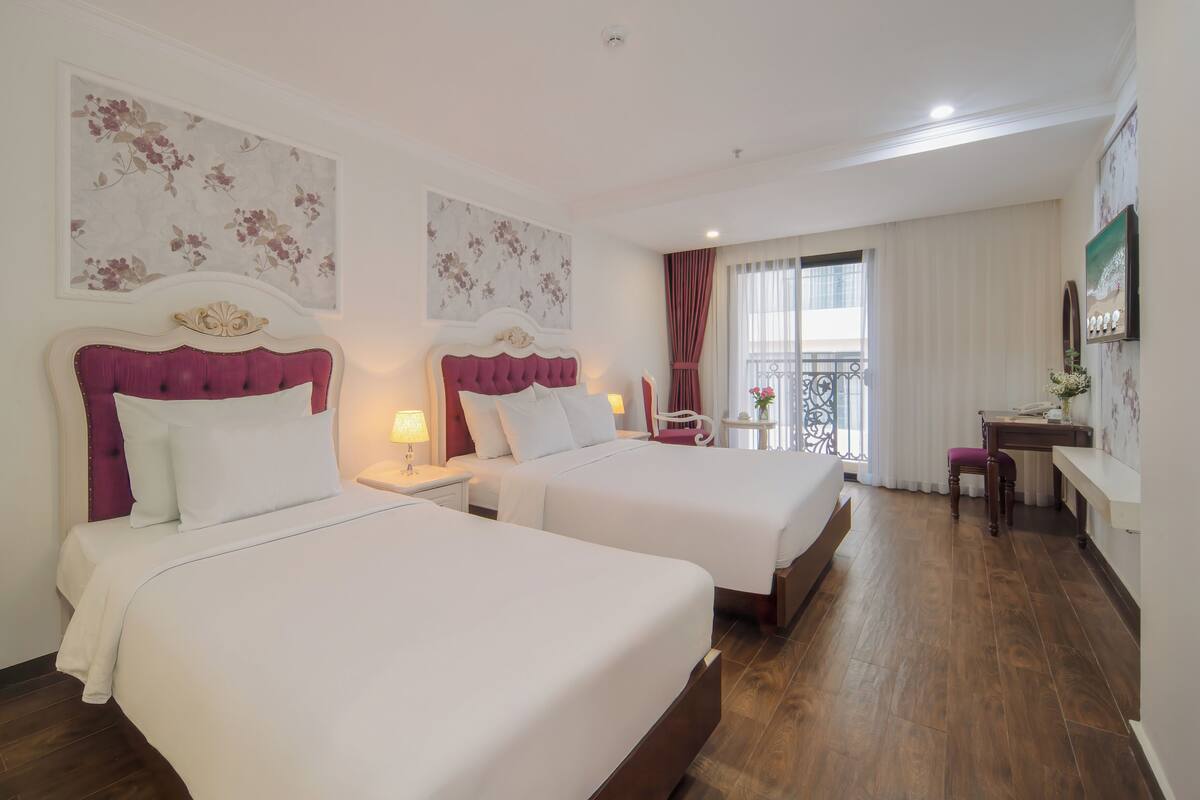A hotel room with two beds, a desk, a chair, and a balcony with a view.