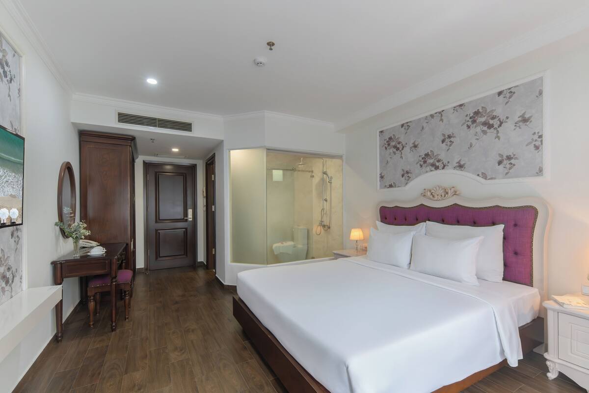 A hotel room with a large bed, a desk with a chair, a vanity, and a shower area.
