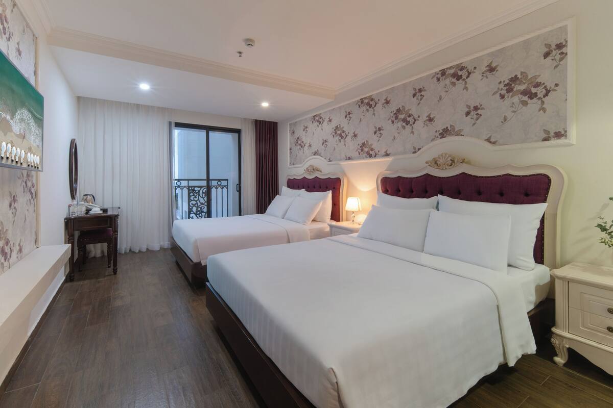 A hotel room with two beds, a bedside table, a nightstand, a window with curtains, and a wall with floral wallpaper.
