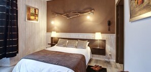 Cama bem arrumada com manta marrom e almofadas, duas luminárias de cabeceira, luminária na parede e um quadro acima da cama.