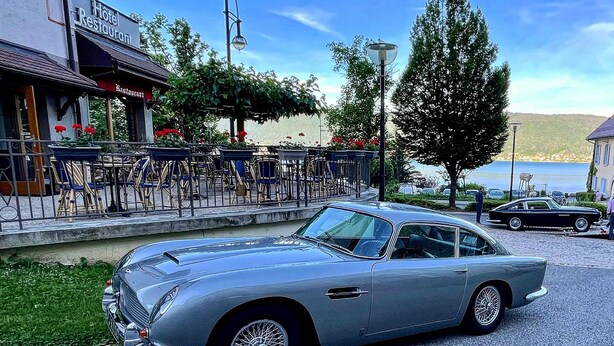 A vintage silver car parked in front of a hotel with outdoor seating and a view of a body of water. - Villa Riva (Veyrier-du-Lac)