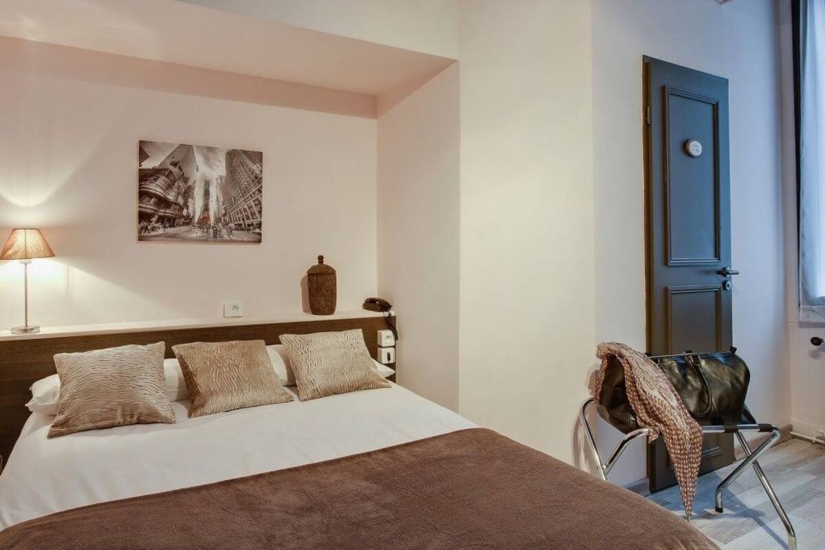 Une chambre d’hôtel avec un lit, une lampe, une chaise, une porte et un tableau accroché au mur.