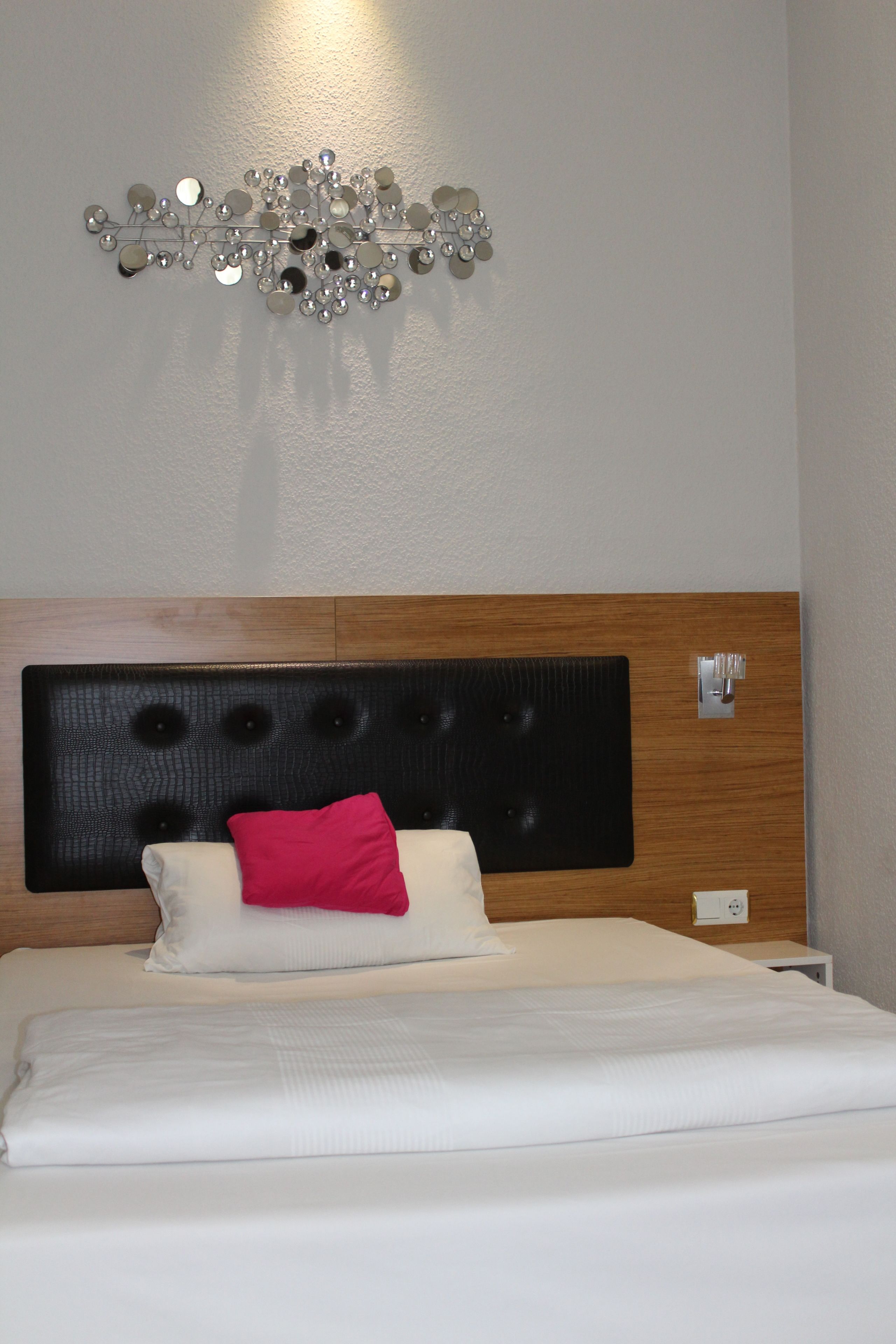 A neatly made bed with white bedding and a single red pillow, a wooden headboard, and a wall-mounted decorative mirror.