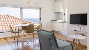 A modern kitchen with white cabinets, a dining area with a table and chairs, and a large window overlooking the sea. - Lofts Azul Pastel (Horta)