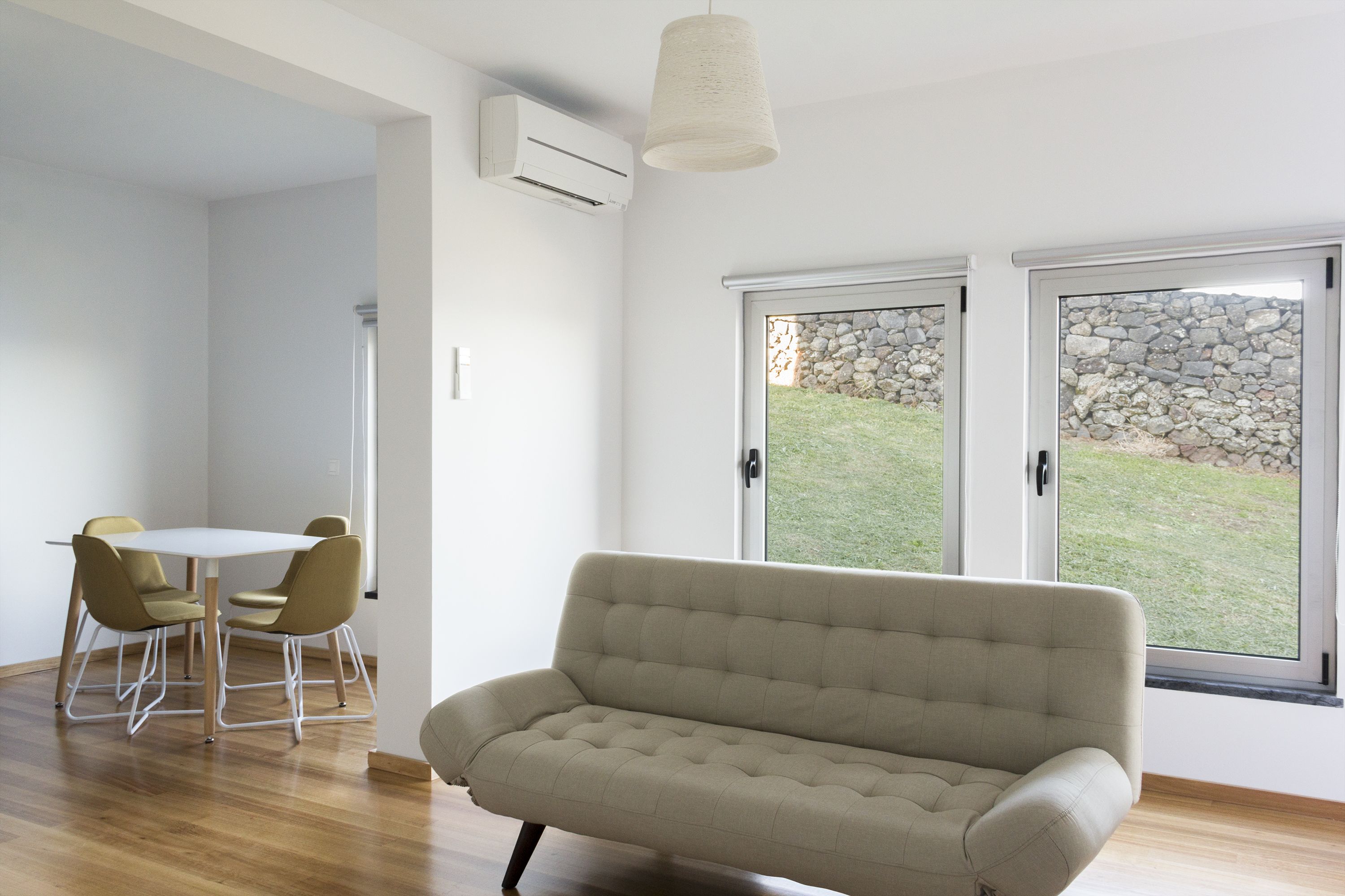 A modern living room with a beige sofa, a dining table with chairs, and large windows with a stone wall view.
